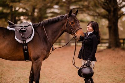 Woman in equestrian attire standing next to a brown horse in an outdoor setting.